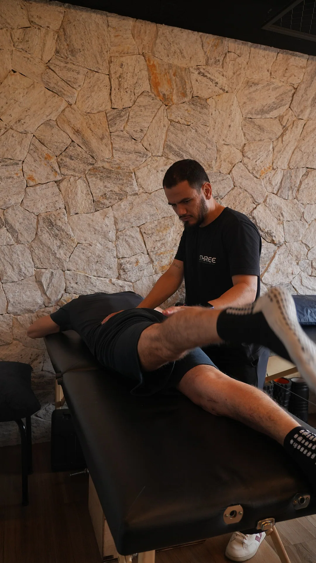 Fisioterapeuta da Three Reabilitação mobilizando o quadril e a perna de um atleta deitado de bruços na maca, com parede de pedra ao fundo